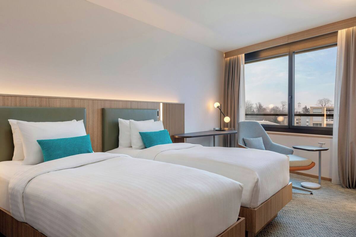 A hotel room with two beds, a desk, a chair, and a view of the city.