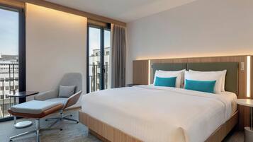 Habitación de hotel con una cama grande, una silla, una mesita y vistas a la ciudad a través de amplios ventanales.