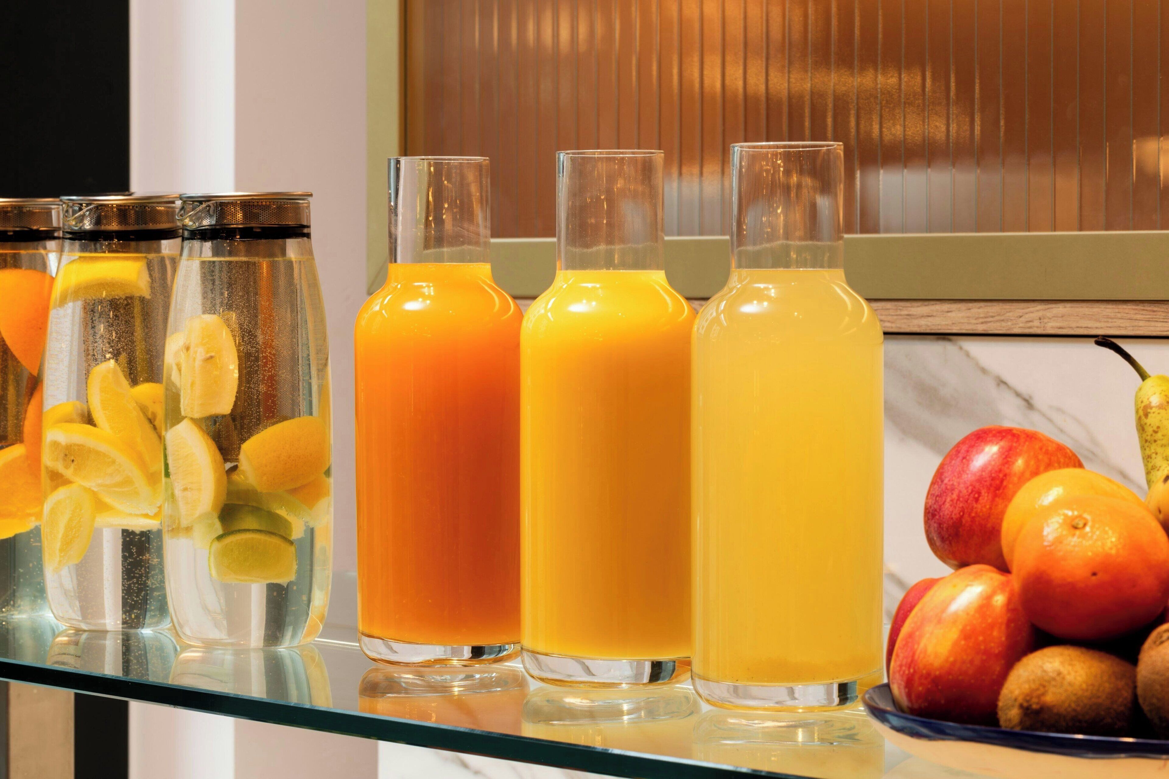 Four glass bottles with orange juice, one with lemon slices, and a bowl of apples and oranges on a glass table.