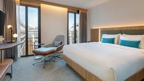 Habitación de hotel con una cama grande, un escritorio con lámpara, una silla y vistas a la ciudad a través de amplias ventanas.
