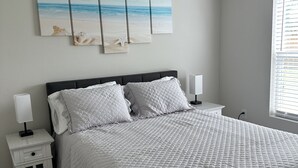 Un dormitorio con una cama grande, una mesita de noche con lámpara y una obra de arte en la pared que muestra una playa.
