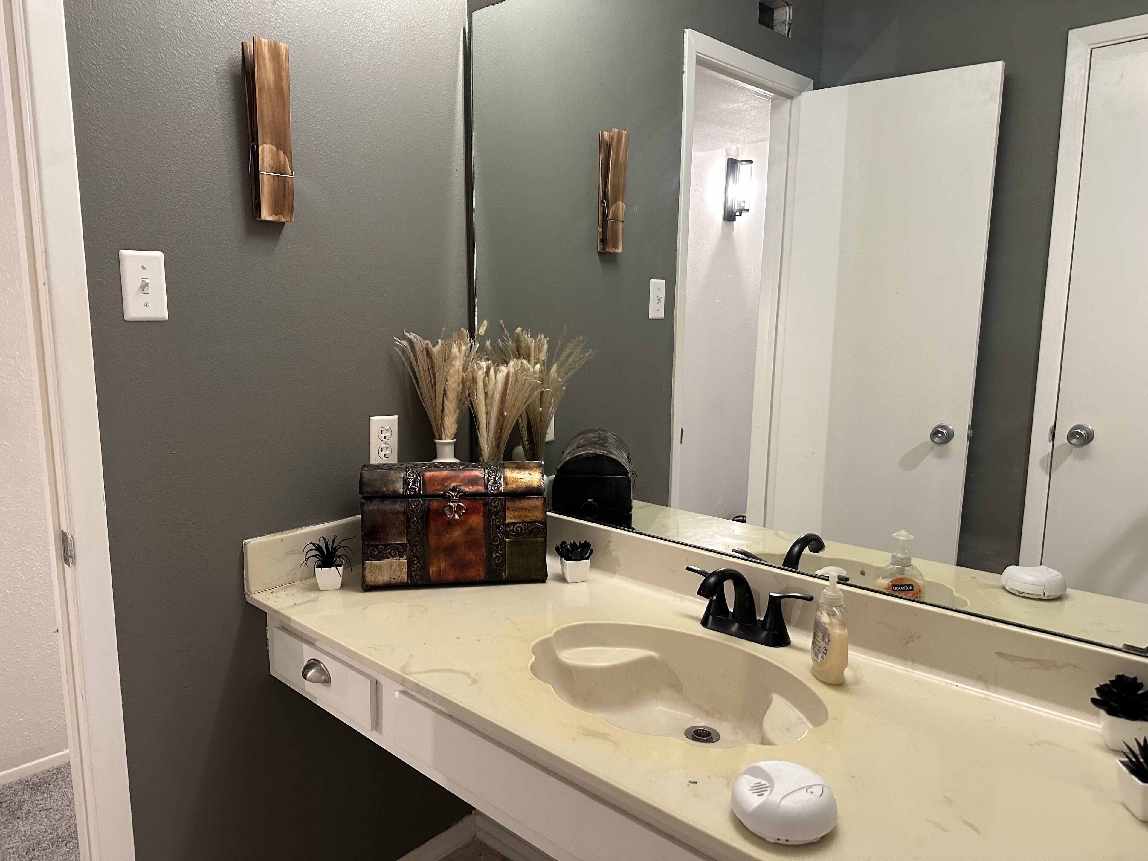Een badkamer met een witte wasbak, een spiegel en een handdoekhouder. Op het aanrecht staan decoratieve items, waaronder een vaas met gedroogde planten.