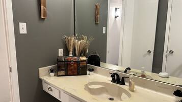 Een badkamer met een witte wasbak, een spiegel en een handdoekhouder. Op het aanrecht staan decoratieve items, waaronder een vaas met gedroogde planten.