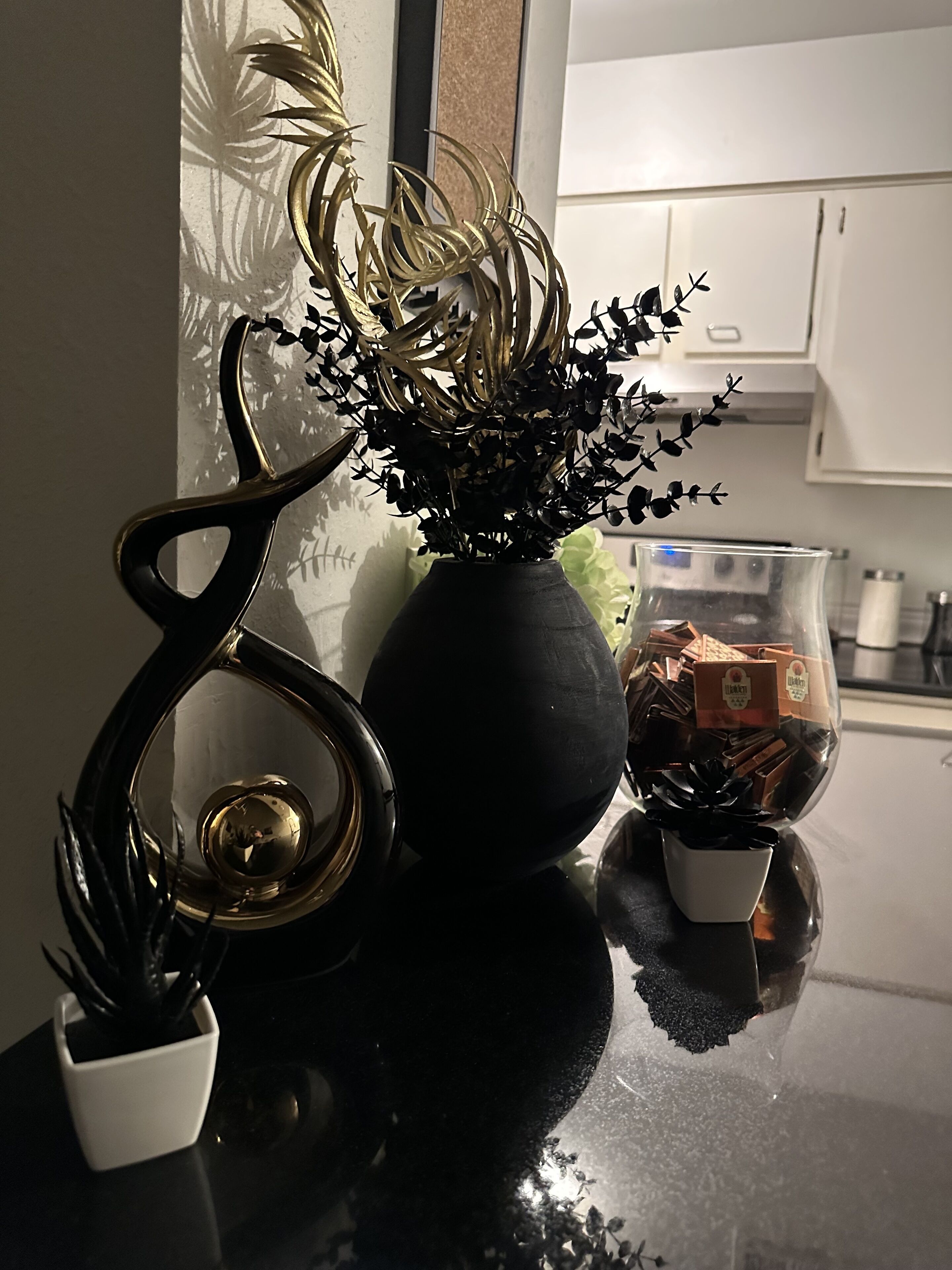 A kitchen counter with a vase of black flowers, a decorative black and gold sculpture, and a glass container with chocolate bars.