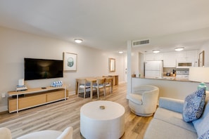 A modern living room with a flat-screen TV, wooden furniture, a dining area, and a kitchen with white cabinets.