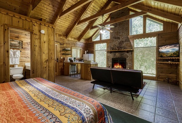 A log cabin interior with a large bed, a fireplace, a stone wall, and wooden beams.