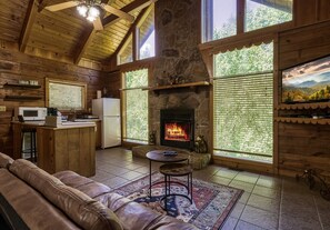 A cozy cabin interior with a stone fireplace, wooden walls, a leather couch, and large windows offering a view of the outdoors.