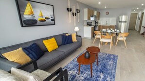 Een moderne woonkamer met een grijze bank, geel en blauw gekleurde kussens, een blauw vloerkleed, en een eethoek met houten stoelen en een kleine tafel.