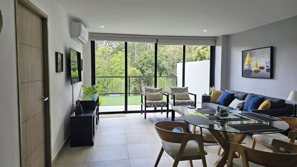 Een moderne woonkamer met een glazen eettafel, een bank en stoelen. Er is een schuifdeur naar een balkon met uitzicht op groen.