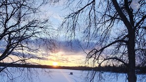 Lumipeitteinen puinen terassi, josta avautuu esteetön näkymä auringonlaskuun jäätyneen järven yllä, ja etualalla näkyvä puu.