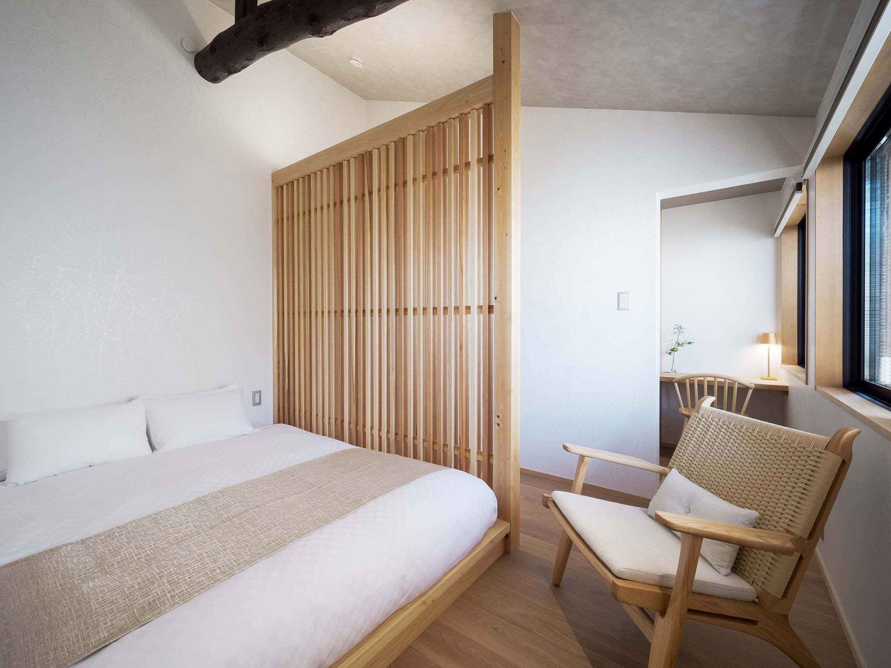 A modern bedroom with a large bed, a wooden partition, a wicker chair, and a desk with a lamp.