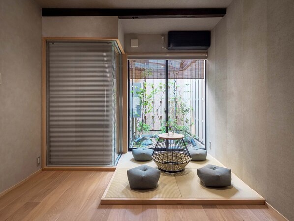 A room with tatami flooring, a sliding door, a small table with cushions, and a view of a garden through a window with a metal grill.