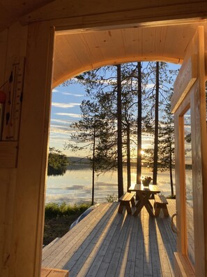 A wooden cabin with a deck overlooking a lake, featuring picnic tables and a view of tall trees. - Cozy Lake Cabin in the Forest (Älvsbyn)