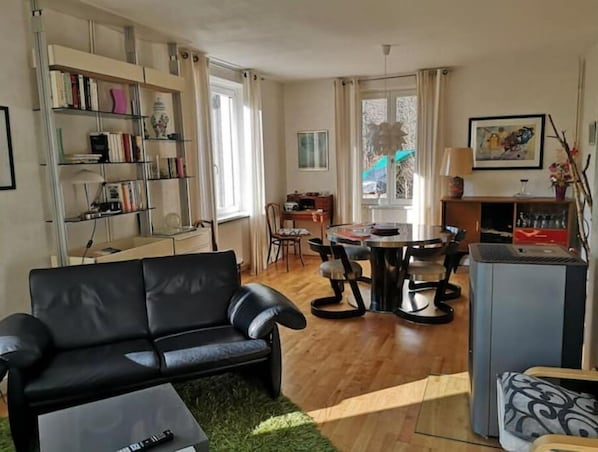 A living room with a black leather sofa, a dining table with chairs, a bookshelf, and a radiator.