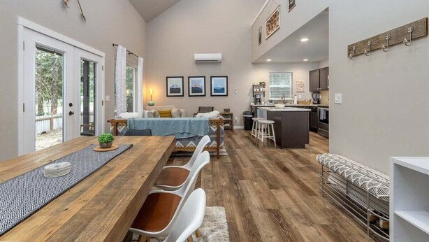 A spacious living area with a wooden dining table, white chairs, and a sofa. There is a kitchen area with a bar counter and stools.