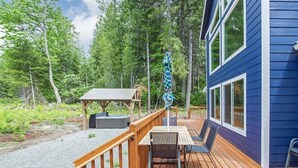 A wooden deck with a table and chairs, a covered seating area, and a building with large windows. - Timbers Edge | Quaint Oasis ~ Hot Tub ~ EV Charger (Packwood)