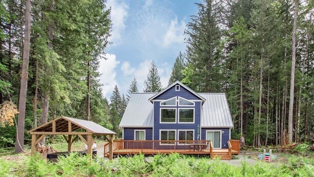 A two-story cabin with a blue exterior and a wooden deck, surrounded by dense forest. - Timbers Edge | Quaint Oasis ~ Hot Tub ~ EV Charger (Packwood)