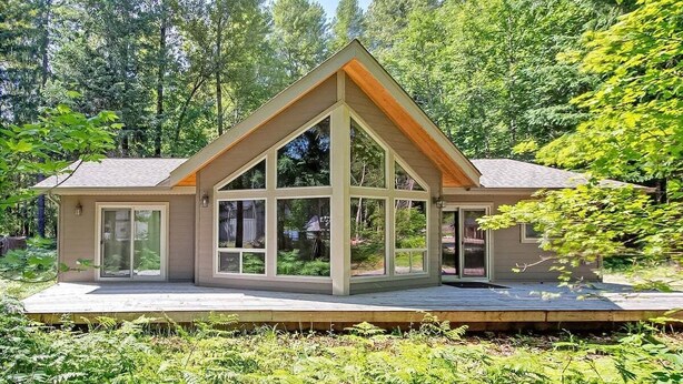 A modern house with a large glass facade, a wooden deck, and a forested backdrop. - Coffee Time Cabin | Hot Tub, Wifi, Grill (Packwood)