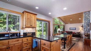 A modern kitchen with wooden cabinets, a central island, and a dining area with a table and chairs. - Coffee Time Cabin | Hot Tub, Wifi, Grill (Packwood)