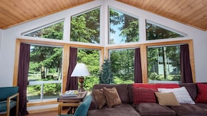 A cozy room with a large window, a brown sofa, a small table, and a lamp. - Coffee Time Cabin | Hot Tub, Wifi, Grill (Packwood)