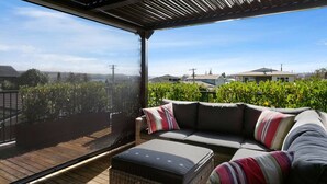 Uma área de estar coberta ao ar livre com sofá e banqueta acolchoados, vista para um vinhedo e céu claro.