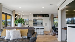 Uma sala de estar moderna com sofá de couro, área de jantar com mesa e cadeiras, cozinha com armários brancos e eletrodomésticos de aço inoxidável, e uma grande janela com vista para o exterior.