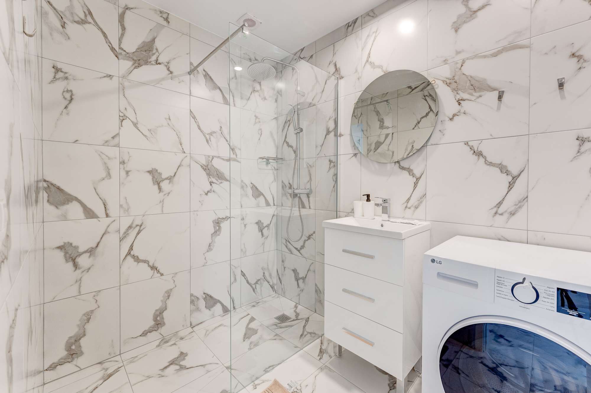 Une salle de bain carrelée de marbre, avec une douche en verre, un lavabo blanc avec un miroir et un lave-linge.