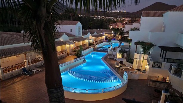 A large, illuminated swimming pool surrounded by white buildings with tiled roofs, set against a mountainous backdrop.