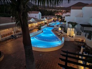 A large, illuminated swimming pool surrounded by white buildings with tiled roofs, set against a mountainous backdrop. - Oasis Golf Spacious Family Duplex (Arona)