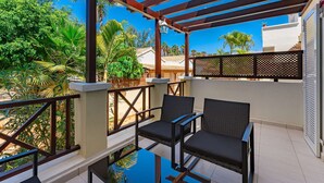 A balcony with two chairs and a glass table, overlooking a garden with palm trees and a white building.