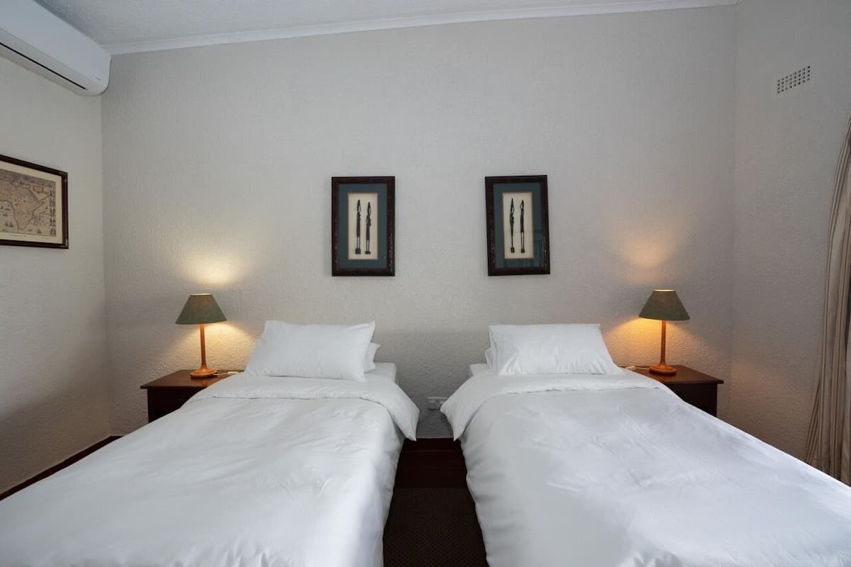 Een hotelkamer met twee eenpersoonsbedden, elk met een nachtkastje en een lamp. Er hangen afbeeldingen in een lijst aan de muur boven de bedden.