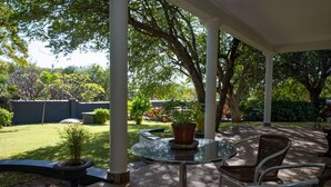 Un patio cubierto con una mesa de vidrio y sillas de mimbre, rodeado de plantas en macetas y árboles.