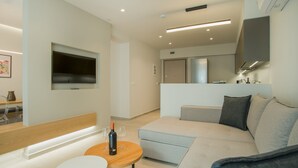 A modern living room with a sofa, a small round table with a bottle and glasses, a TV mounted on the wall, and a kitchen area in the background. - CELINE LUXURY APARTMENTS & SUITES (Agios Nikolaos)