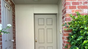Eine beigefarbene Tür mit vier Feldern, eine Fußmatte mit der Aufschrift "Welcome" und ein kleiner, grüner Busch zur Rechten.