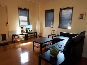 A living room with a sofa, coffee table, and TV. There are three windows with blinds, a framed picture on the wall, and a plant on the coffee table. - 2BR Fashionable Townhouse Flemington / Showgrounds / Melbourne (Kensington)