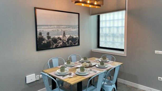 Un coin repas avec une table dressée pour quatre personnes, une photographie encadrée représentant un paysage côtier accrochée au mur et une fenêtre avec des stores.
