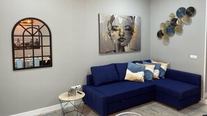 Un salon avec un canapé d’angle bleu, une petite table ronde, un tableau abstrait accroché au mur et un élément décoratif en métal fixé au mur.