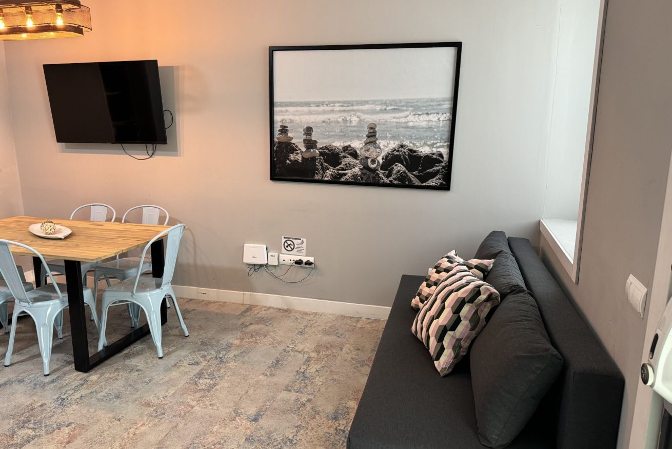 A room with a dining table, chairs, a sofa, a TV mounted on the wall, and a framed picture of a beach scene.