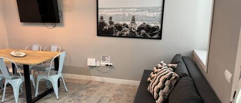 A room with a dining table, chairs, a sofa, a TV mounted on the wall, and a framed picture of a beach scene.