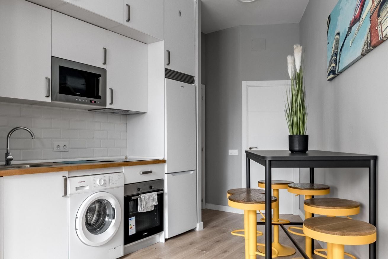 A modern kitchen with a washing machine, microwave, and refrigerator. There is a dining area with a table and stools.