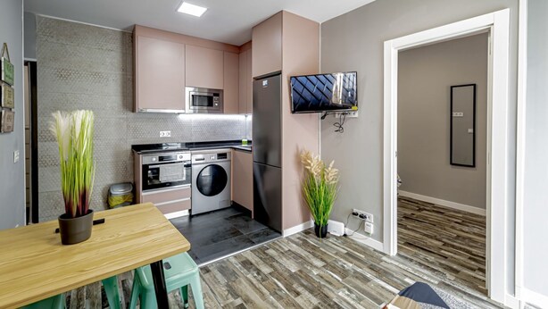 Et kompakt køkken med moderne hårde hvidevarer, et spiseområde med bord og stole, og en opholdsstue med et fjernsyn.