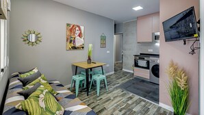 En kompakt opholdsstue med sofa, et lille spisebord, et tekøkken og et fjernsyn ophængt på væggen.