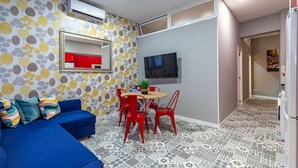 Ein kompakter Wohnbereich mit blauem Sofa, roten Sesseln, einem kleinen Esstisch, einem Fernseher und einem Fußboden mit Muster.