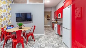 Ein kompakter Wohnbereich mit Essbereich, ausgestattet mit einem runden Tisch und roten Stühlen, einer Küche mit roten Schränken und einem weißen Kühlschrank, sowie einem an der Wand montierten Flachbildfernseher.