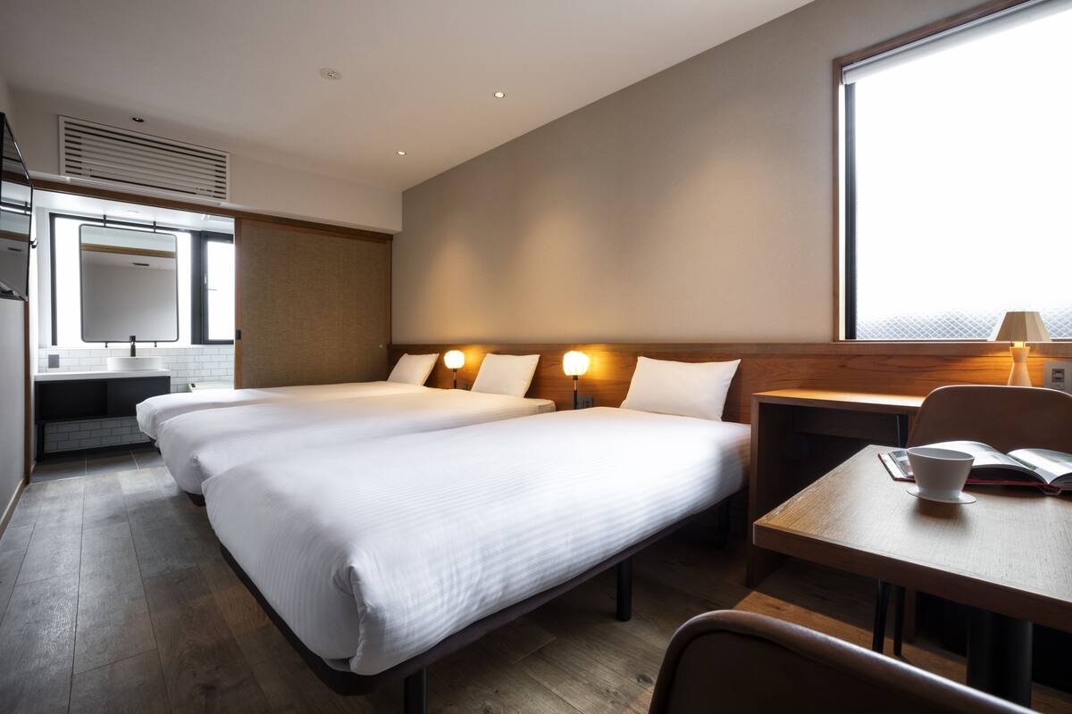 A hotel room with two beds, a desk, a chair, a window, and a bathroom with a mirror and sink.