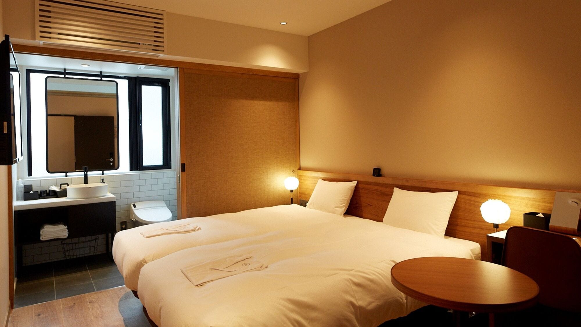 A hotel room with a double bed, a bedside table, a round table, a sink, and a mirror.