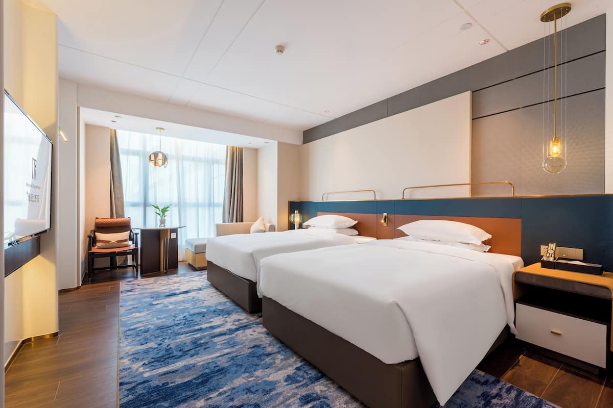 A hotel room with two beds, a TV, a desk, and a large window.