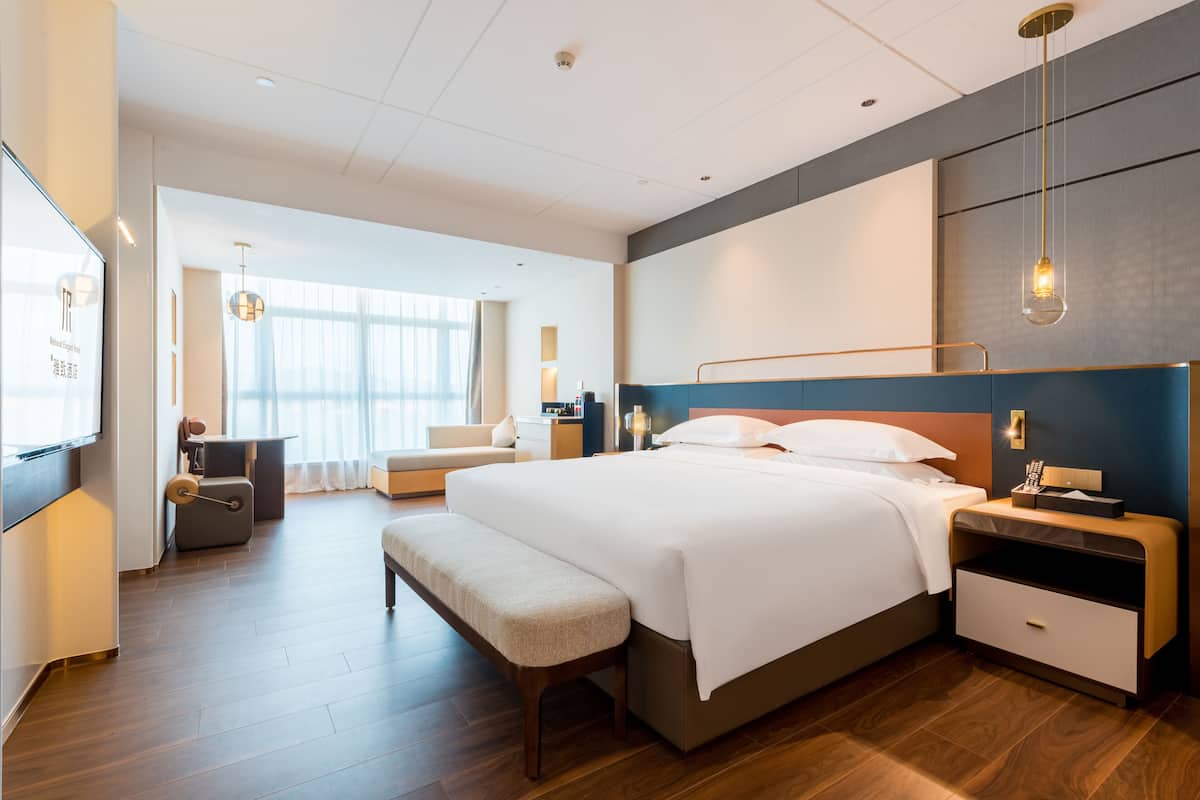 A modern hotel room with a large bed, bedside tables, a bench, and a seating area with a sofa and armchair.