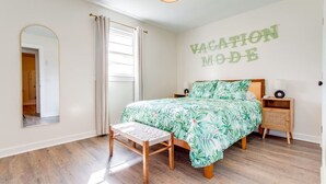 Un dormitorio con cama, una banca, una mesita de noche, un espejo y una pared con el texto 'VACATION MODE'.