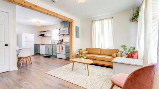 Una sala moderna con un sofá, una mesa de centro y un sillón. Se observa una cocina al fondo con armarios y electrodomésticos.